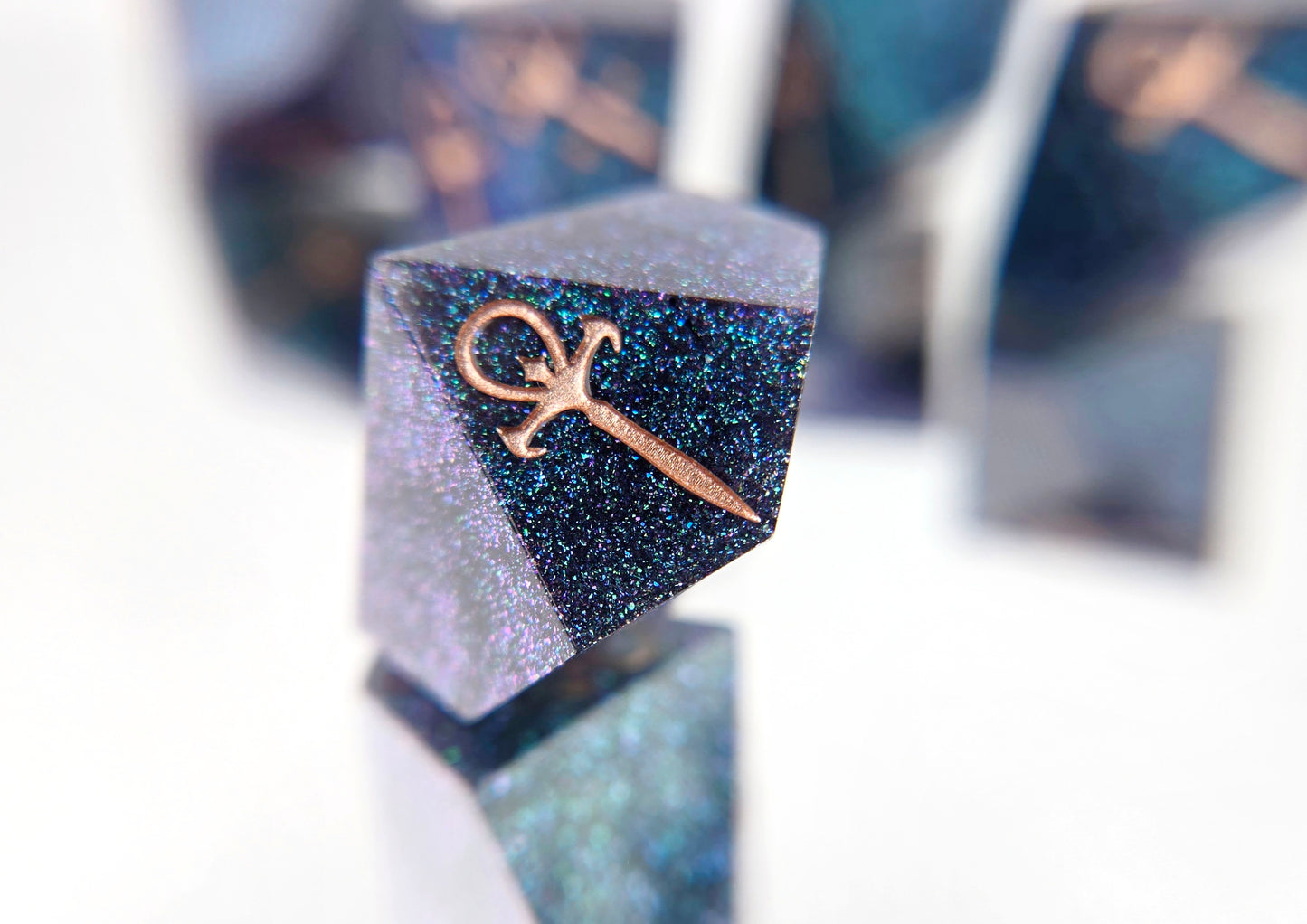 A closeup of a Vampire the Masquerade d10 focusing on the ankh symbol. The dice are densely packed blue and green colorshift glitter with rose gold inking for the symbols.