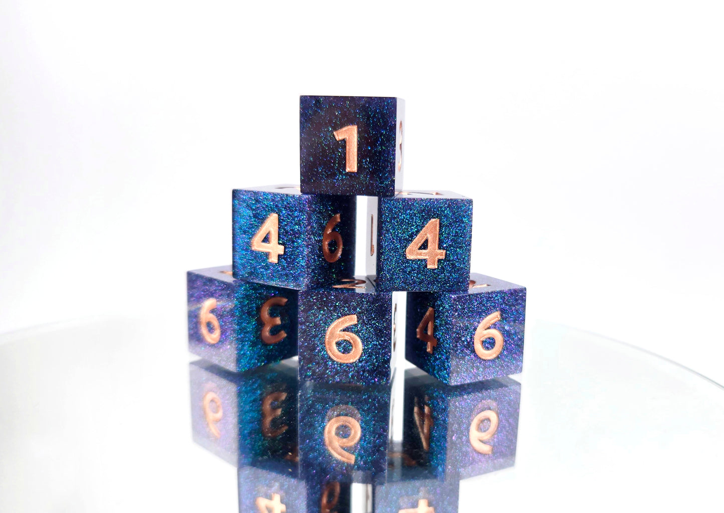 A stack of 6 d6 dice. The dice are densely packed blue and green colorshift glitter with rose gold inking for the symbols.